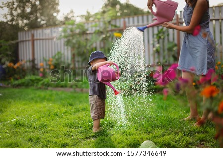 Mom and boy child water the garden together, Mom’s little gardener assistant, taking care of children and garden. Mother watering her son from watering can, take care of trees and plants, wet child