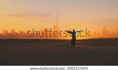 Arabic woman weared in traditional UAE dress - abayain rising her hands on the sunset at a desert with Dubai city silhouette on the background.