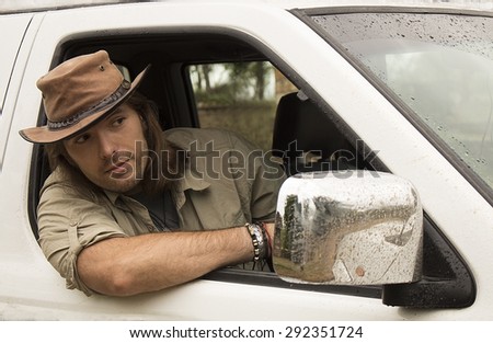 Handsome young man with long hair in brown cowboy hat driving white 4x4 car after rain. Safari style.