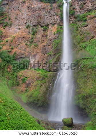 Narrow frothy waterfall bordered by red rock, moss, and green plants falls into a lagoon
