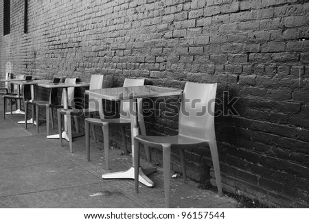 Black and white image of brushed aluminum tables and chairs on a sidewalk against a black brick wall