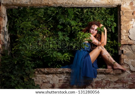 Wild woman with white body henna tattoos posing on gained by bushes window of abandoned building