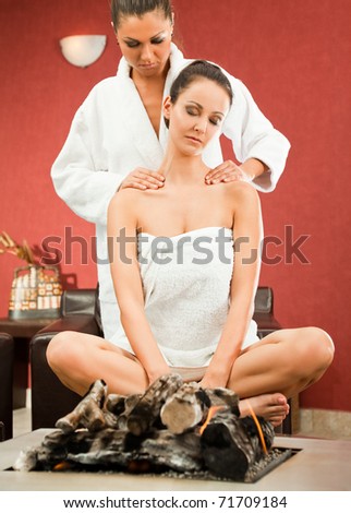 Young female sitting behind fireplace with legs crossed, receiving shoulder massage by woman