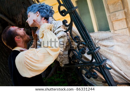Male vampire holding woman's neck, both dressed in medieval costumes