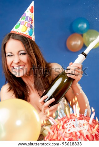 Happy excited female with party hat sitting behind birthday cake, popping up champagne
