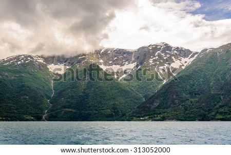 Panoramic view on the three mountain peaks with snow caps with the blue surface of the water, magic grey clouds on the sky, Sognefjord, Norway