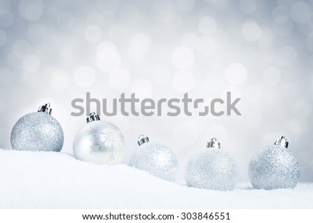 A row of silver Christmas baubles on snow with defocused silver and white lights in the background. Shallow depth of field.