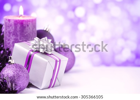 Purple and silver Christmas baubles, a gift and a candle in front of defocused purple and white lights.