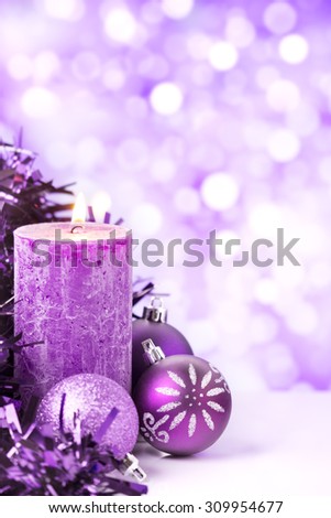 Purple and silver Christmas baubles and a candle in front of defocused purple and white lights.