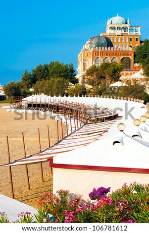 Beach cabanas at a luxury Italian art deco hotel.  Cabanas are on the sand in the foreground, the grand hotel is in the background.  Shot against a bright blue sky.