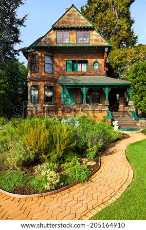 Victorian house with natural brown wood shingles and painted green trim. In front is a flower and herb garden with a winding brick path.