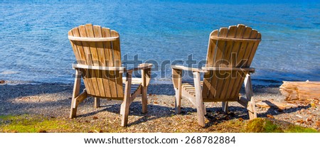 Two wooden adirondack chairs on the shore of a lake facing the water. Viewed from the back.