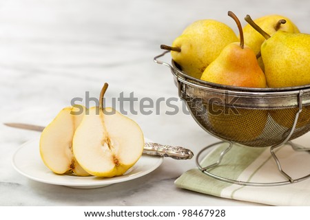 Yellow pears in a vintage metal strainer, with a sliced pear on a white plate with a vintage silver knife. Shot on a white marble counter top.