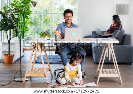 Photo of Mixed race family sharing time in living room. Caucasian father using notebook computer to work and half-Thai playing and painting under desk while Asian mother with laptop working her job on sofa.