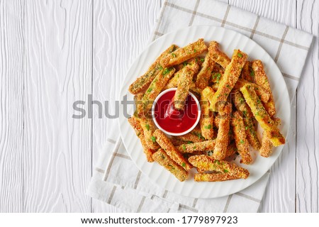 crunchy zucchini sticks breaded with panko breadcrumbs, parmesan cheese, spices on a white plate with ketchup on a wooden table, view from above, flat lay, free space