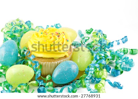 A large yellow cupcake with sprinkles surrounded by blue and green Easter Eggs and curly ribbons on a white background with copy space