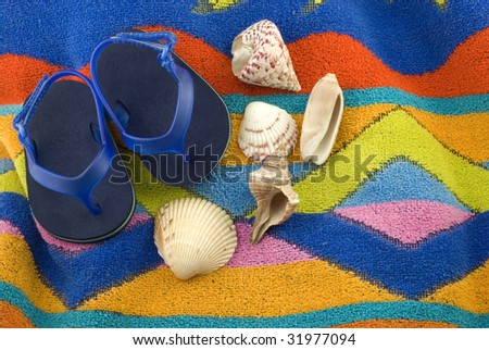 A pair of tiny blue infant sandals on a colorful beach towel with a collection of seashells, horizontal with copy space