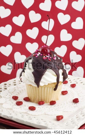 A Valentine sundae cupcake with white frosting, fudge, topped with a cherry and sprinkles, heart background, selective focus, vertical