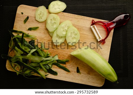 Wooden cutting board with peeled and cut cucumber, vegetable peeler and cucumber peelings on a black background