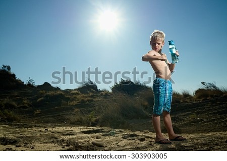 A young 7 year old boy, in the summer heat, having a blue, translucent, plastic water bottle filled with water  in his hand, looking at his muscles
