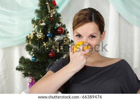 The young beautiful woman sits near a christmas fur-tree and eat an orange