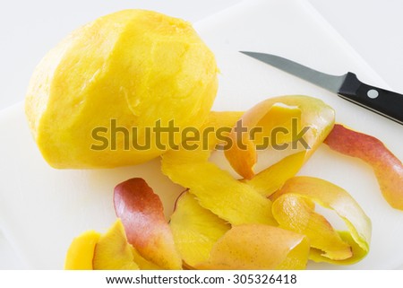 A mango that has been peeled.  The peel is in a pile next to the whole, ripe mango and the paring knife used to peel it is in the background.