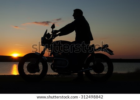 Motor-biker in silhouette at sunset  infron of lake man sitting on  motorbike  both hands on handlebar