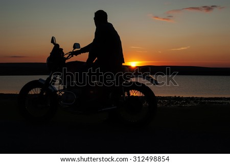 Motorbiker in silhouette at sunset  by lake man on motorcyclist one hand on handlebar