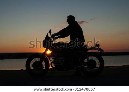Motorcyclist  in silhouette at sunset by lake man sitting on  motorbike  both hands on handlebar