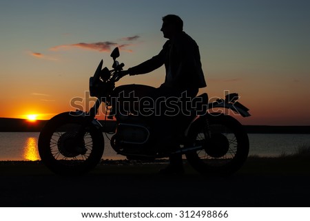 Motorbiker in silhouette at sunset by lake man on motorbike one hand on handlebar