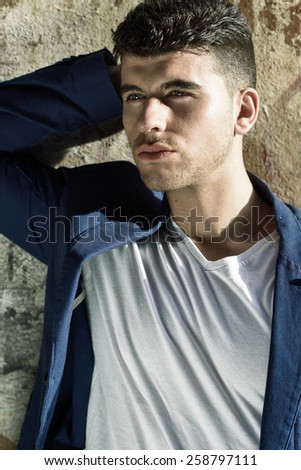 Close-up portrait of a young man with blue eyes posing near a wall. Model of fashion in urban background wearing white t-shirt and blue jacket
