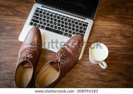a pair of brown leather shoes placing on a laptop computer and there is a cup of milk beside the computer, classic wooden table, selective focus, foreign keyboard