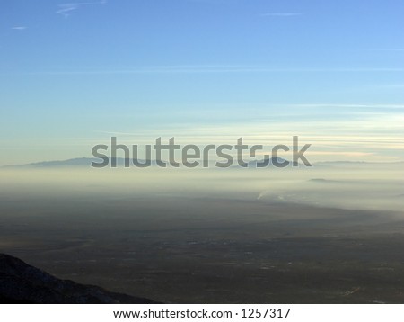 Two mountains poke above the New Mexico desert haze with whispy clouds in the blue sky above.  Just below the right mountain, you can see smoke from a burning fire.