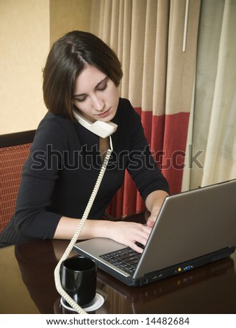 A businesswoman talks on the phone while working on her laptop computer in a hotel room during a business trip.