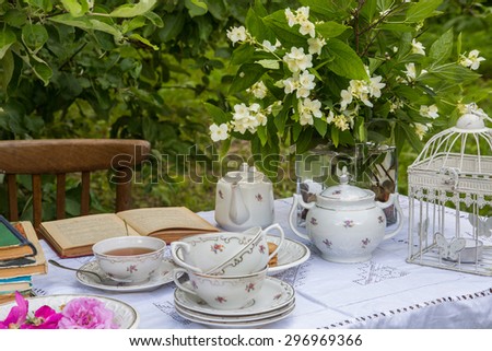 A summer tea party. A cup of green tea and empty cups, a teapot, a sugar bowl, cookies, books, white cage and flowers on the table in the garden.