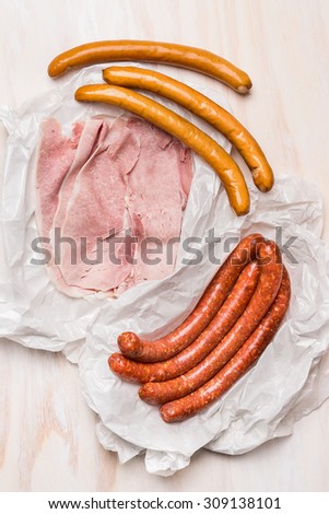 different long sausages and ham in a crumpled paper packaging on rustic wooden background, top view