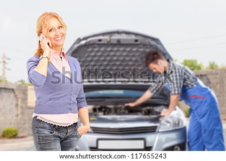 Smiling careless female talking on a mobile phone while in the background mechanic is checking her car