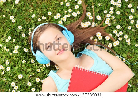 Young female with notebook listening music and lying on a grass with daisies
