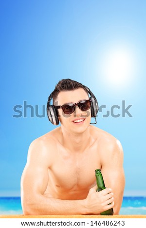 Smiling guy with headphones and sunglasses drinking cold beer, on beach next to a sea