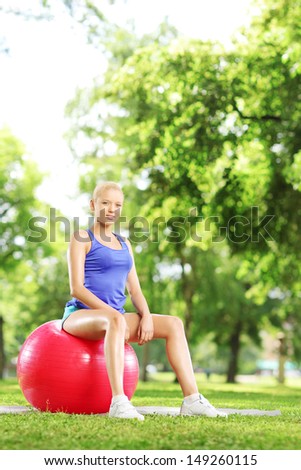 Young blond female sitting on a pilates ball and looking at camera in park