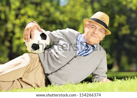 Senior man with hat lying on a green grass and holding a soccer ball in a park