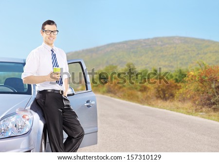 Smiling young man on his automobile relaxing and drinking coffee on an open road, shot with a tilt and shift lens
