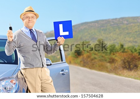 Senior man posing on his car, holding a L sign and car key after having his driver\'s licence on an open road