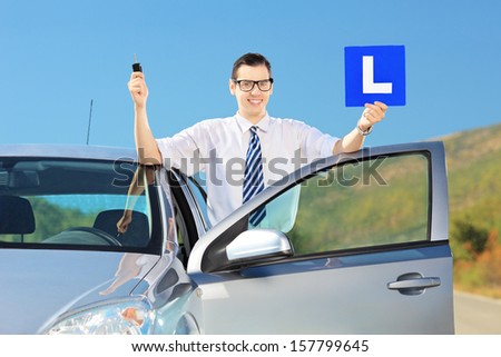 Happy young man posing near his car, holding a L sign and car key after having his driver\'s licence on a road
