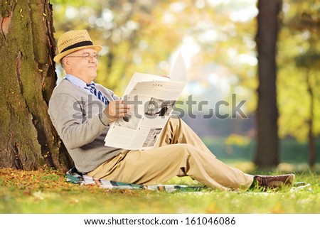 Smiling senior gentleman seated on a grass reading a newspaper in a park at autumn