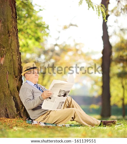 Smiling senior gentleman seated on a grass reading a newspaper in a park at autumn, shot with a tilt and shift lens