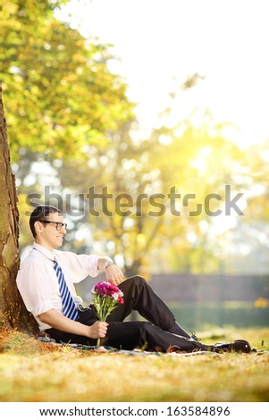 Handsome guy with a bunch of flowers sitting ona  grass and checking the time, in a park