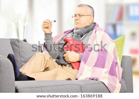 Sick mature man on a sofa with a hot-water bottle looking at thermometer at home