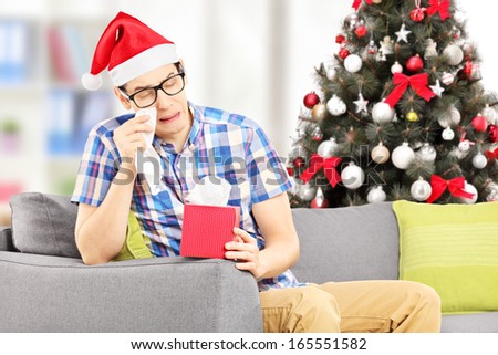 Sad young male seated on a sofa wiping his eyes from crying with christmas tree in the background