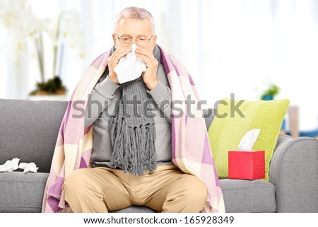 Sick senior man covered with blanket sitting on a sofa and blowing his nose at home